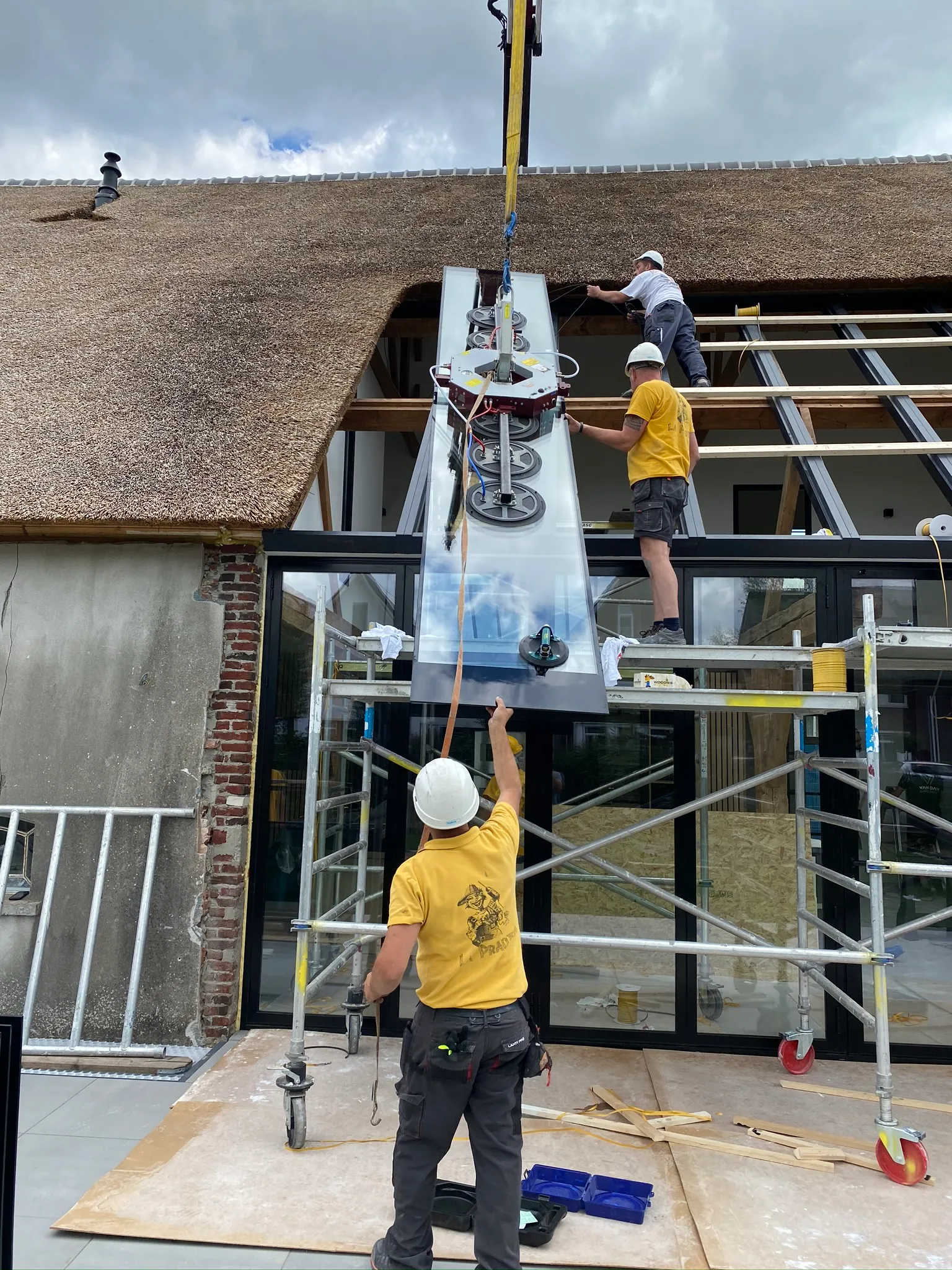Medewerkers in La Pradera-shirts plaatsen met een hijskraan een groot glaspaneel bij de restauratie van een boerderij